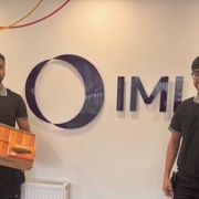 Prizewinners of the £1000 prize draw holding their box of products in front of an IMI sign in the reception of the Blackburn IMI Heatmiser site.