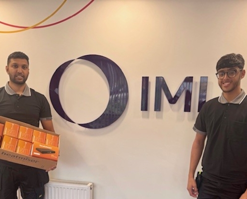Prizewinners of the £1000 prize draw holding their box of products in front of an IMI sign in the reception of the Blackburn IMI Heatmiser site.
